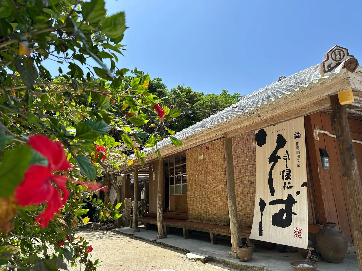 赤い花の奥に建つ今帰仁そばの古民家の店舗。軒先に大きなのれんがかかる
